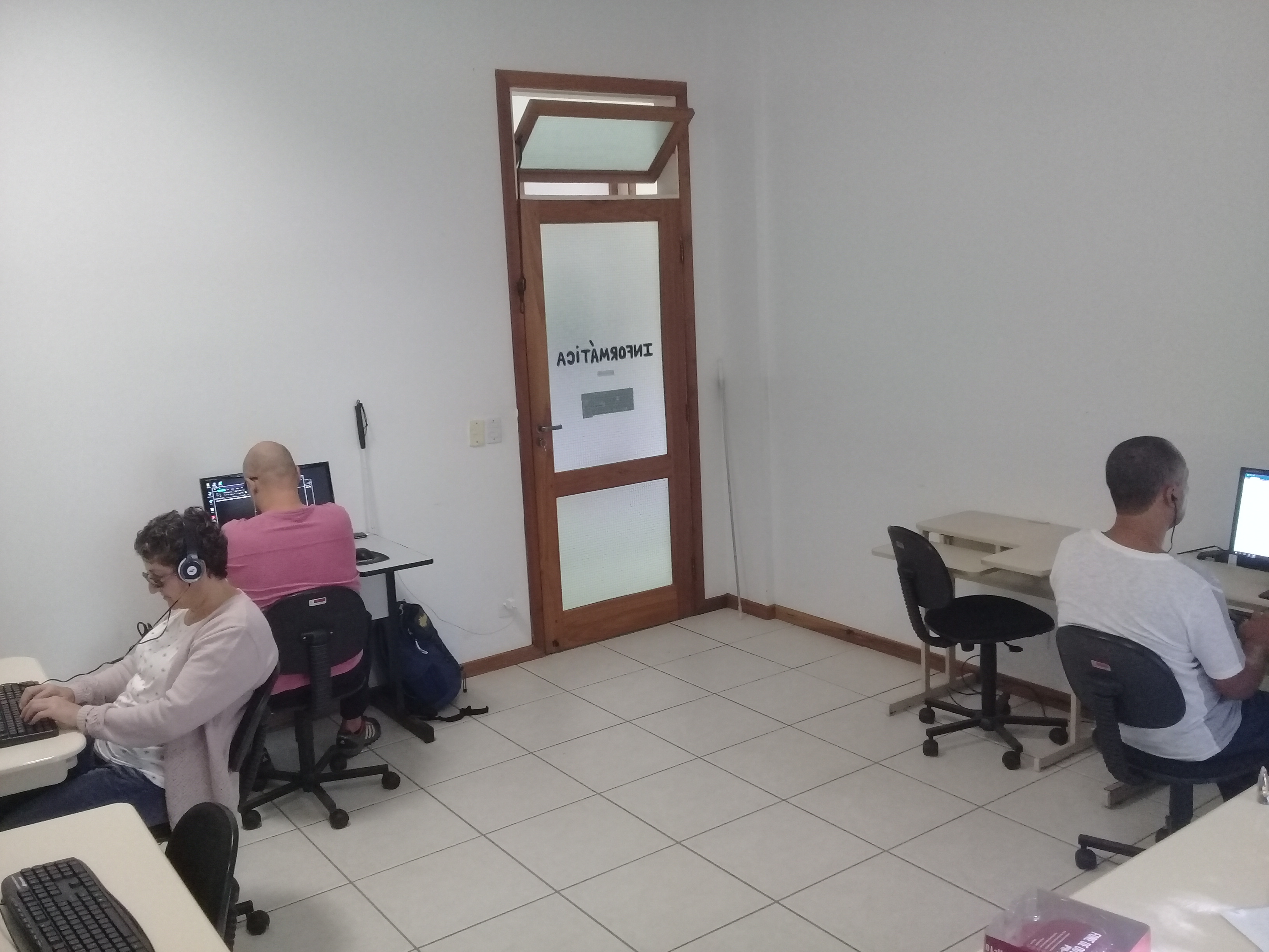 na imagem aparecem os alunos Félix, Rodrigo e Zenilde sentados em frente ao computador. Os três alunos usam fones de ouvido e estão praticando as técnicas de digitação no Word, com o auxílio do leitor de tela.