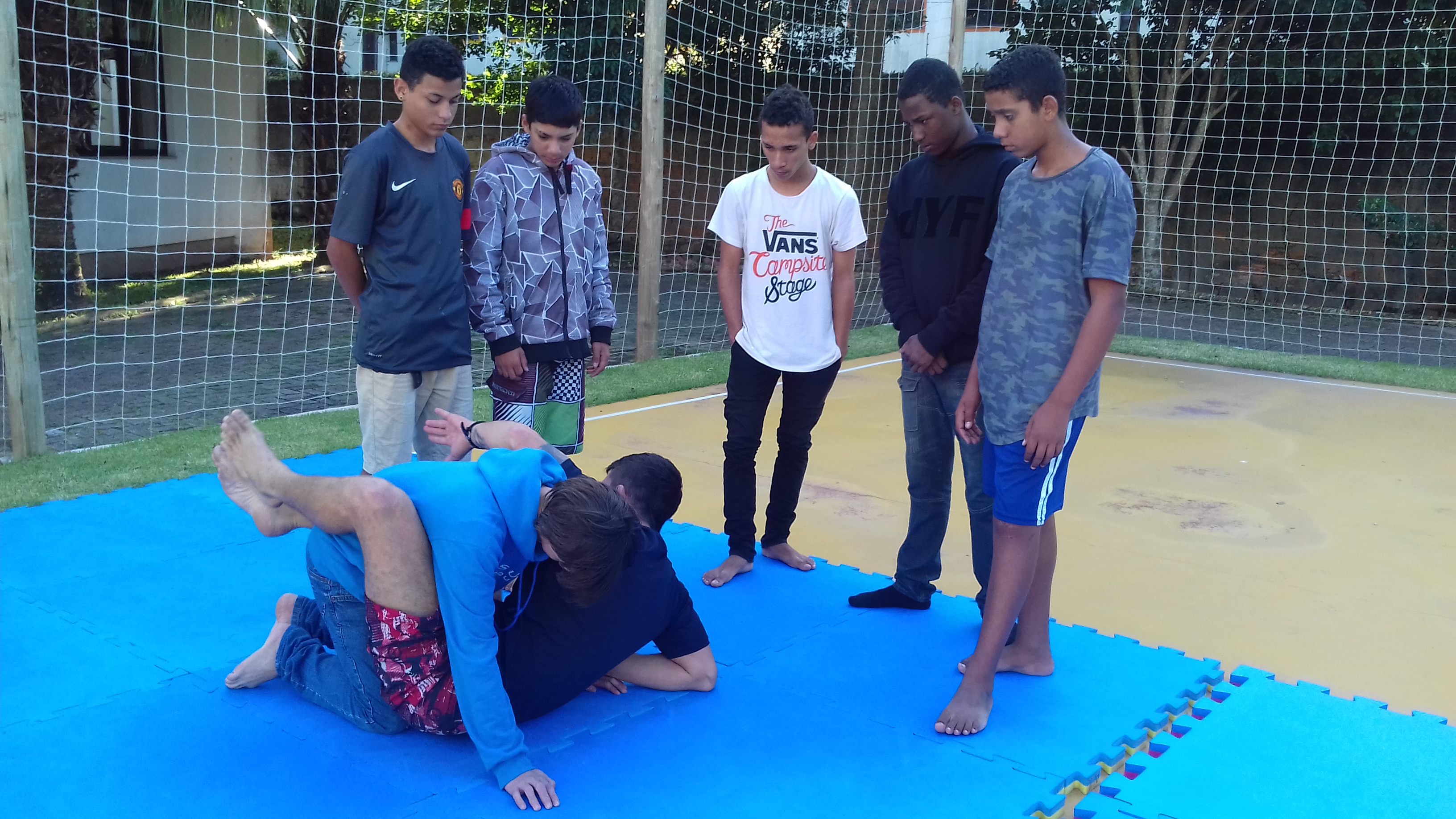 Na imagem tatames na quadra de esportes da ACIC Profº Fernado executando movimentos de chave de ombro Jiu Jitsu com um aluno e os outros 5 alunos em pé observando, alunos adolescentes.